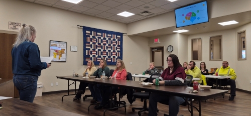 classroom with instructor to the far left, and attendees sitting at long, fold up tables, listening to the instructor